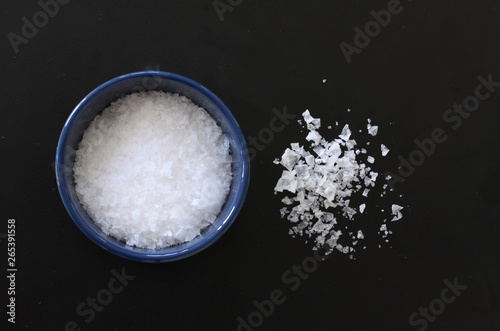 Flake salt lying in a blue bowl and scattered around on a black background. Top view image.