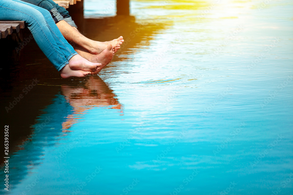 Romantic couple man and woman are sitting on a dock with barefoot feet dangling in pond water. Sunny weather. Happiness, vacation, summer, concept. Copy space for text.