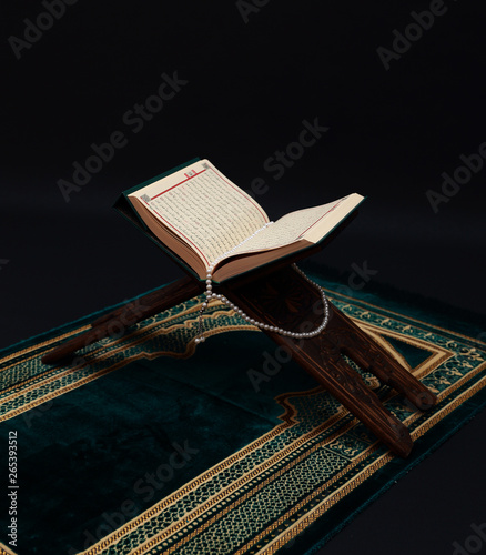 Islamic Holy Book Quran on wood carving rahle with rosary beads and prayer rug on black background. Kuran the holy book of Muslims. Ramadan concept.