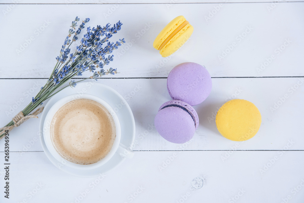 Fototapeta premium Sweet delicious violet and yellow macarons and cup of latte or americano and branch of fragrant lavender on white wooden background.