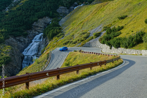 Light blue car races through a hairpin turn running past glassy mountain stream.