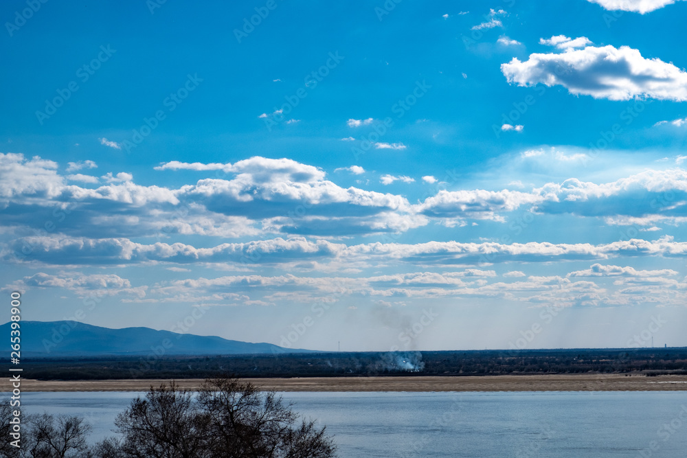 View of the Amur river against the blue sky with white beautiful clouds. Bright spring sun. Russia, Khabarovsk. View of the Amur river against the blue sky with white beautiful clouds. Bright spring sun. Russia, Khabarovsk.