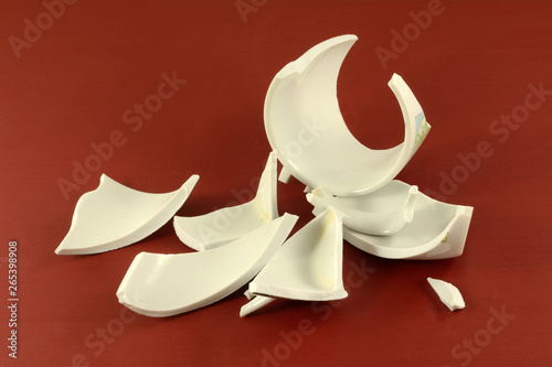 Broken pieces of white drinking mug on red wooden background as symbol of brokenness in world