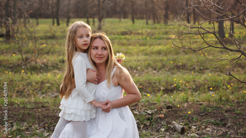 Family portrait of a single mother hugging her daughter. They are posing in white dresses against the backdrop of a spring meadow. Mothers Day. The girl has a bouquet of forest tulips in her hand
