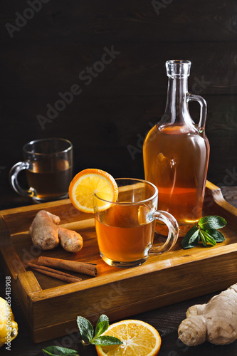 Homemade Kombucha in a glass decanter and glasses on a wooden tray with ginger, mint and lemon on a dark background. This is a healthy natural fermented sweet and sour drink originally from China
