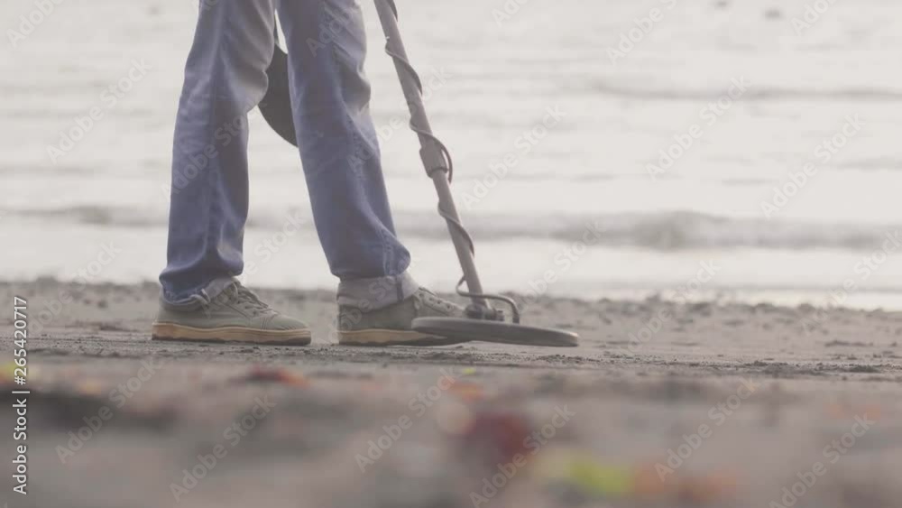 Man is working on the beach holding metal detector finding metal in the sand on the coastline at sunrise.