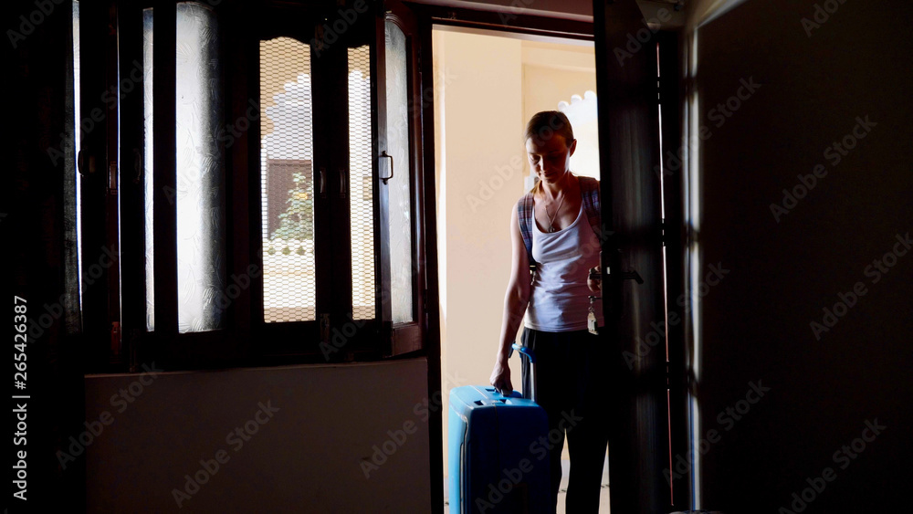 Silhouette of young woman in the hotel with a suitcase opens the door and passes to the dark room. View from the inside.