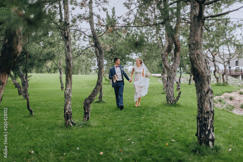 Stylish groom in shirt and beautiful blonde bride are walking, holding hands, in the forest, against the background of green grass and trees. Wedding portrait of smiling and cheerful newlyweds.