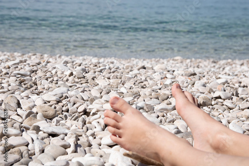 Child's feet laying on white pebble beach with blue sea water backgroung photo