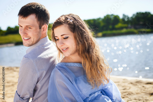 young lovely couple resting on beach, handsome guy and cute girl smiling and sitting back to back on coast river, happy family concept