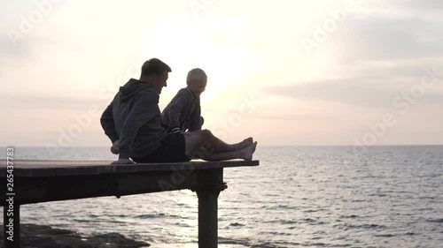 The family watches a beautiful sunset. A man and a boy sit on a wooden bridge on a rock by the sea and watch the sun set into the sea. Son sits in the arms of the Pope and shows his hand in the sun