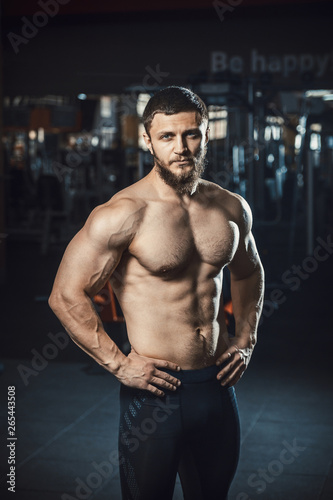 Good looking young man bodybuilder with beard standing posing in front of the mirror at the gym darkened slogan background . Athlete showing straining veins on hands and bubble guts vertical