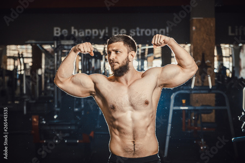 Good looking young man bodybuilder posing in front of the mirror shows big biceps at the gym darkened slogan background. Athlete showing straining veins on hands bubble guts