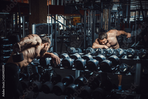 Good looking young man bodybuilder doing triceps kickback lifting dumbbells in front of the mirror looking at himself. athlete making exercises for the triceps near dumbbell racks
