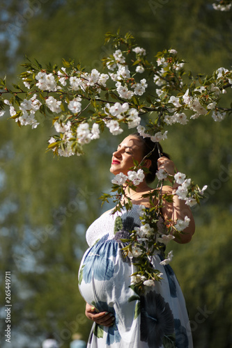 Happy young soon to be mother mom - Young traveler pregnant woman enjoys her leisure free time in a park with blossoming sakura cherry trees wearing a summer light long dress with flower pattern