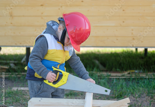 boy in hard hat with a handsaw sawing a wooden Board. young carpenter at work near the new wooden house