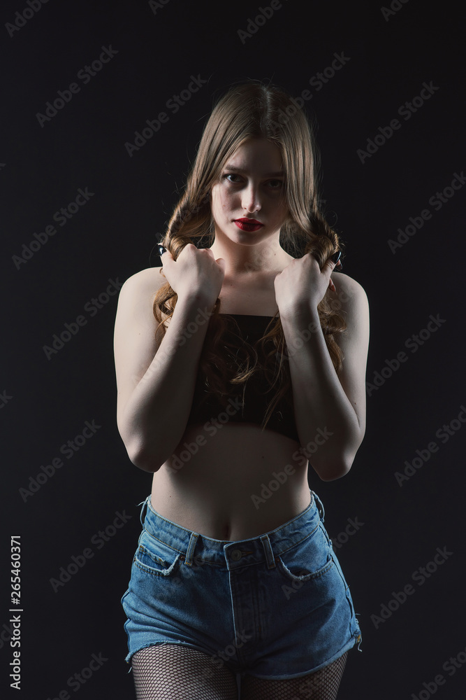 Young beautiful, slender, passionate brown-haired woman with bright red lips and long hair in a black top and denim shorts on a dark background Young beautiful, slender, passionate brown-haired woman with bright red lips and long hair in a black top and denim shorts on a dark background