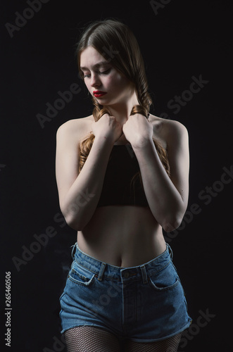 Young beautiful, slender, passionate brown-haired woman with bright red lips and long hair in a black top and denim shorts on a dark background