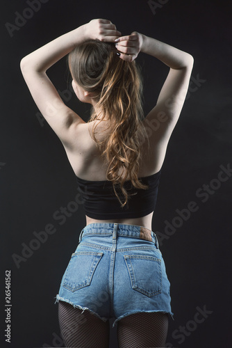 Young beautiful, slender, passionate brown-haired woman with bright red lips and long hair in a black top and denim shorts on a dark background