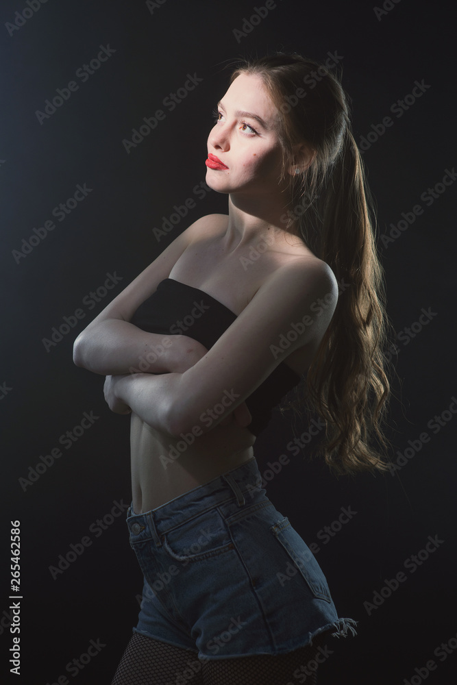 Young beautiful, slender, passionate brown-haired woman with bright red lips and long hair in a black top and denim shorts on a dark background Young beautiful, slender, passionate brown-haired woman with bright red lips and long hair in a black top and denim shorts on a dark background