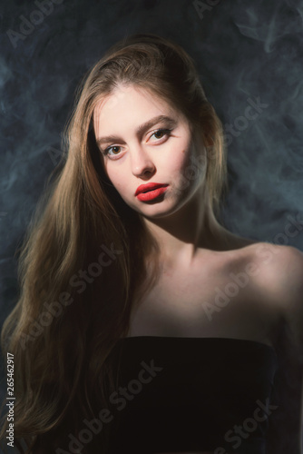 Young beautiful, slender, passionate brown-haired woman with bright red lips and long hair in a black top and denim shorts on a dark background