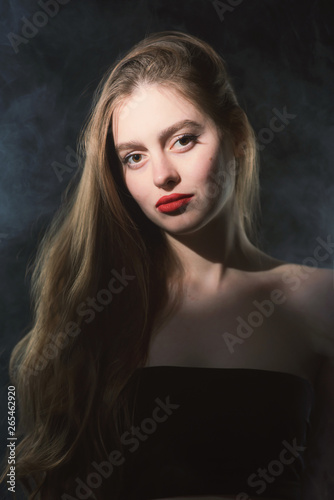 Young beautiful, slender, passionate brown-haired woman with bright red lips and long hair in a black top and denim shorts on a dark background
