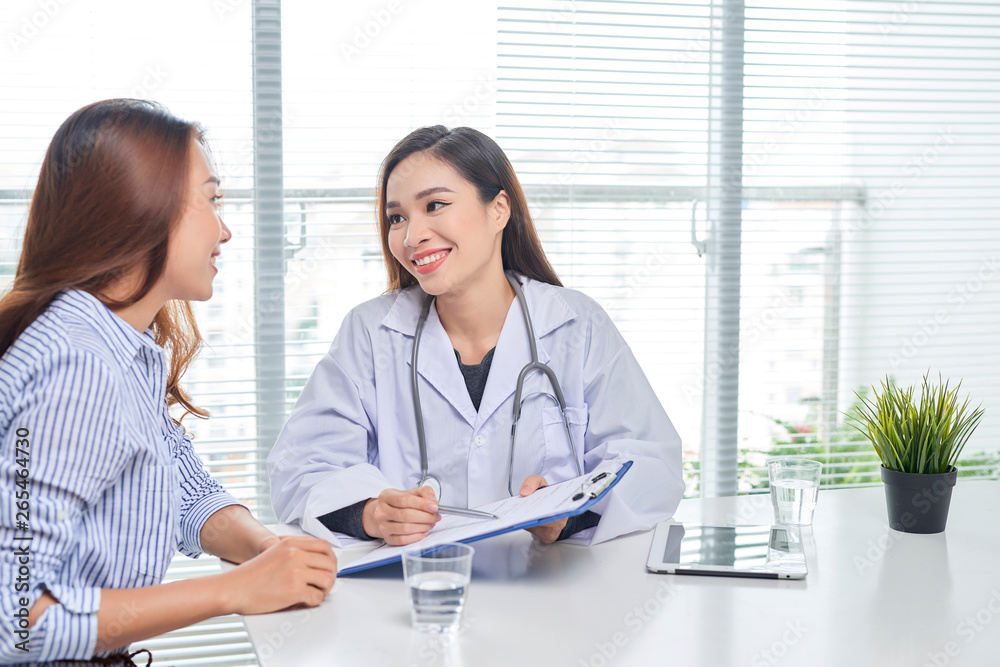 Female doctor talks to female patient in hospital office while writing on the patients health record on the table. Healthcare and medical service. Female doctor talks to female patient in hospital office while writing on the patients health record on the table. Healthcare and medical service.