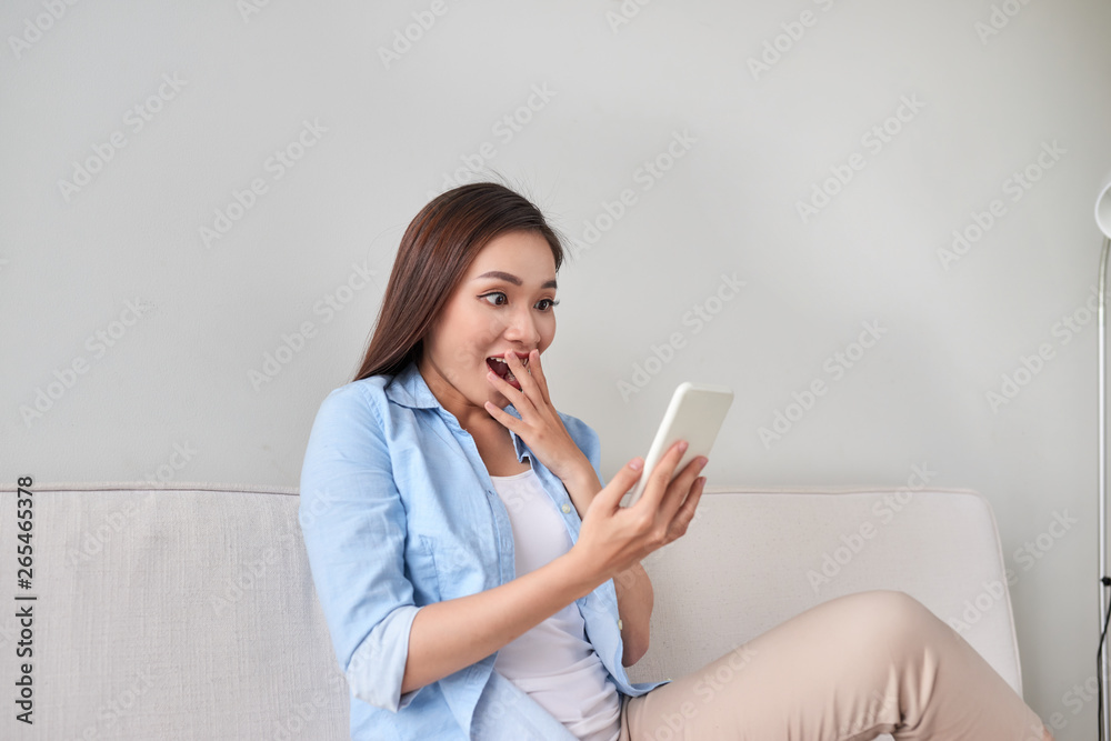 Excited woman reading text on a phone lying on a couch in the living room at home Excited woman reading text on a phone lying on a couch in the living room at home