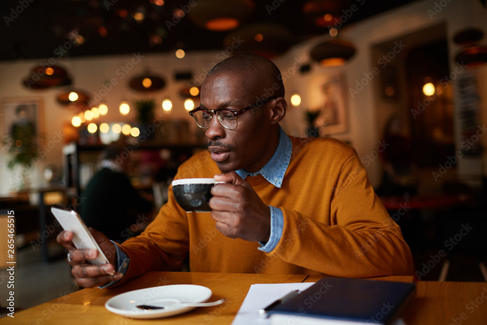 Warm toned portrait of contemporary African-American man using smartphone sitting at table in coffee shop, copy space Warm toned portrait of contemporary African-American man using smartphone sitting at table in coffee shop, copy space