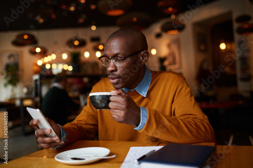 Warm toned portrait of contemporary African-American man using smartphone sitting at table in coffee shop, copy space