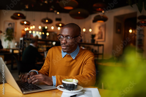Warm toned portrait of contemporary African-American man using laptop sitting at table in coffee shop, copy space