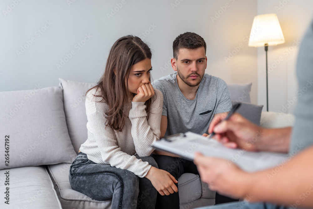 Unhappy couple at therapy session with man talking to therapist in therapists office. Sad couple listening to a marriage counselor during a therapy sitting on a sofa at home Unhappy couple at therapy session with man talking to therapist in therapists office. Sad couple listening to a marriage counselor during a therapy sitting on a sofa at home