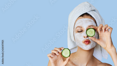 Beautiful young woman with facial mask on her face holding slices of cucumber. Skin care and treatment, spa, natural beauty and cosmetology concept. photo