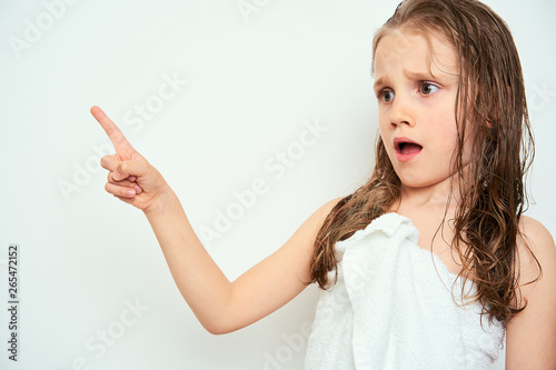 Little preschool girl with wet hair photographed against white background wrapped in white towel pointing with finger having dazed facial expression