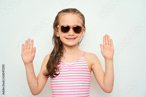 Smiling little preschool girl with wet hair photographed against white background wearing swimsuit and sunglasses demonstrates surrender gesture