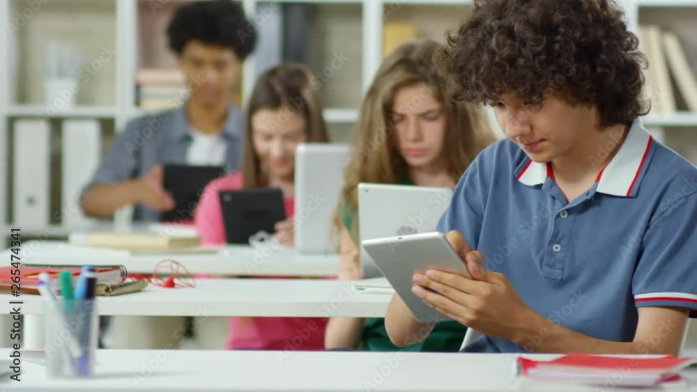 Tracking right of young Caucasian people sitting at school desks and using tablets