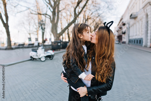 Portrait of two lovely adorable sisters standing in the middle of the street in morning on blur background. Long-haired young woman with trendy hair accessory holding daughter and gently kissing her.