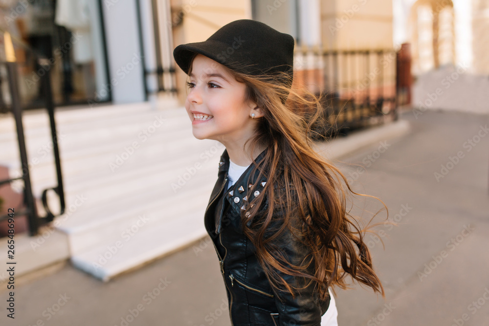 Happy little girl running down the street with dark hair waving and looking away. Portrait of playful brunette girlie wearing black hat and stylish jacket jumping on city background in holidays Happy little girl running down the street with dark hair waving and looking away. Portrait of playful brunette girlie wearing black hat and stylish jacket jumping on city background in holidays