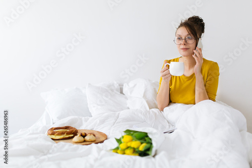 Young woman having breakfast in bed and using mobile phone