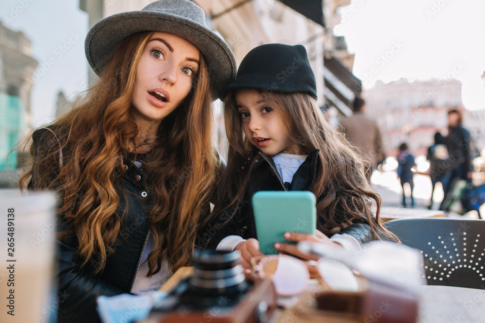 Brown-haired woman in leather jacket and brunette girl in trendy hat posing with surprised face expression waiting for call from dad. Portrait of cute young mom and lovely astonished daughter