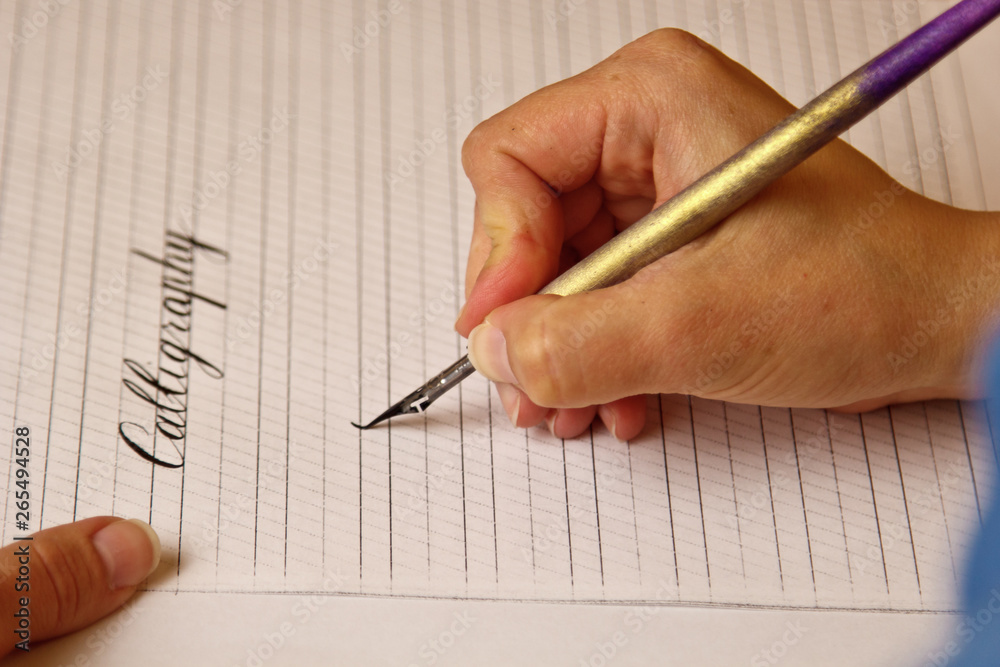 female hand writes with the inky pen the word calligraphy on a paper sheet with stripes. stationery on desk close up top view. spelling lessons and caligraphy exercises. Template, layout, background female hand writes with the inky pen the word calligraphy on a paper sheet with stripes. stationery on desk close up top view. spelling lessons and caligraphy exercises. Template, layout, background