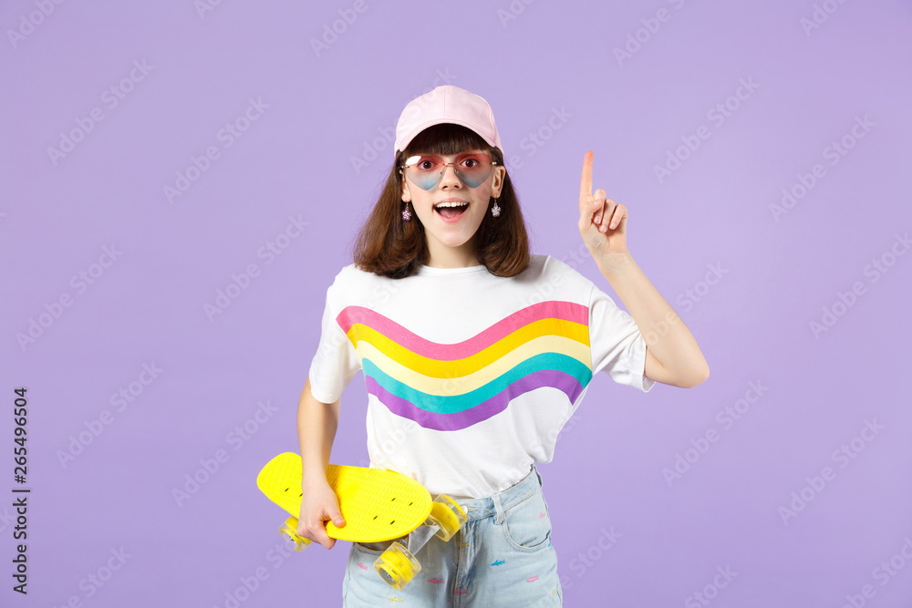Excited teen girl in vivid clothes, eyeglasses with skateboard holding index finger up with great new idea isolated on violet background. People sincere emotions lifestyle concept. Mock up copy space. Excited teen girl in vivid clothes, eyeglasses with skateboard holding index finger up with great new idea isolated on violet background. People sincere emotions lifestyle concept. Mock up copy space.