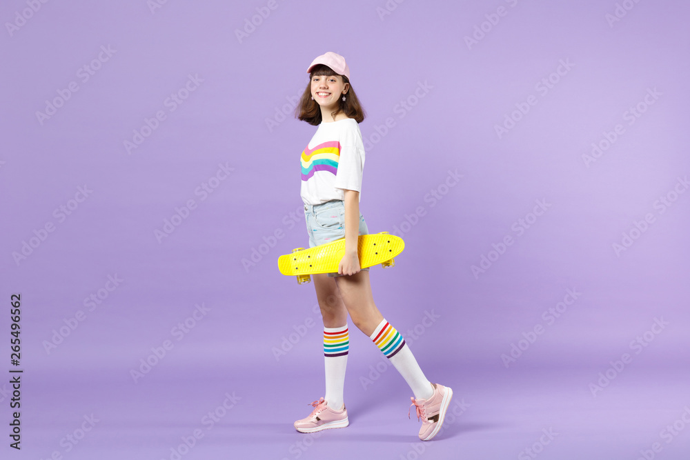 Φωτογραφία Side view of funny teen girl in vivid clothes holding yellow skateboard, looking camera isolated on violet pastel background in studio