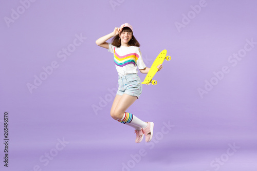 Portrait of smiling teen girl in vivid clothes holding yellow skateboard, having fun, jumping isolated on violet pastel wall background. People sincere emotions, lifestyle concept. Mock up copy space.