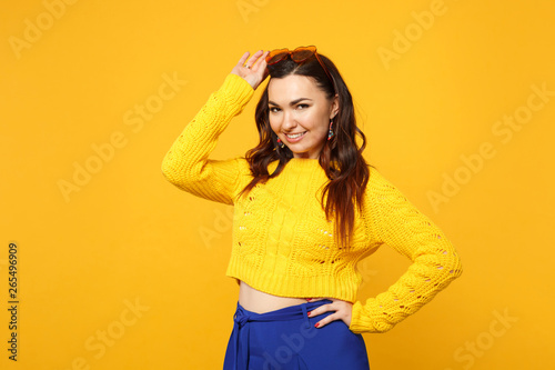 Portrait of pretty young woman in sweater, blue trousers, heart glasses standing, looking camera isolated on yellow orange background. People sincere emotions, lifestyle concept. Mock up copy space.