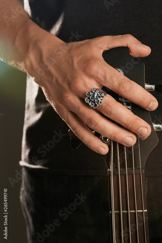 Cropped shot of man's hand, touching headstock of guitar. The guy is wearing massive signet-ring with blacking in view of skulls. The man is wearing black clothes, posing against the dark background.