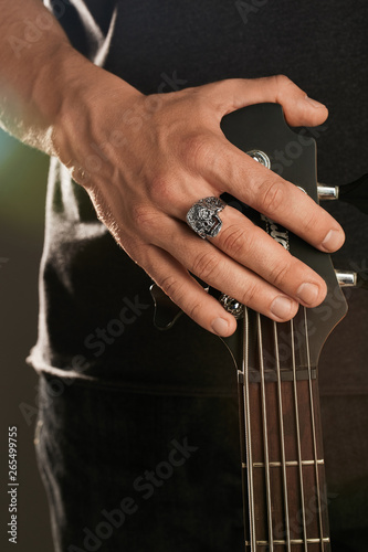 Cropped shot of man's hand, touching headstock of guitar. The guy is wearing signet-ring in the view of skull with opened jaws. The man is wearing black clothes, posing against the dark background.
