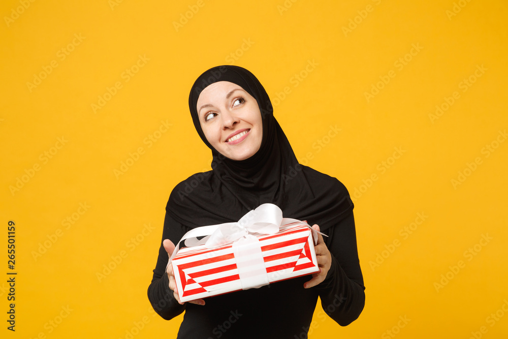 Smiling young arabian muslim woman in hijab black clothes hold in hand present box with gift isolated on yellow wall background studio portrait. People religious lifestyle concept. Mock up copy space. Smiling young arabian muslim woman in hijab black clothes hold in hand present box with gift isolated on yellow wall background studio portrait. People religious lifestyle concept. Mock up copy space.