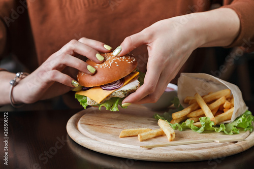 The girl is holding a delicious Burger - a fresh sesame bun  a juicy cutlet  lettuce leaves  onions  a juicy tomato and a slice of cheese. French fries and greens on a wooden plate