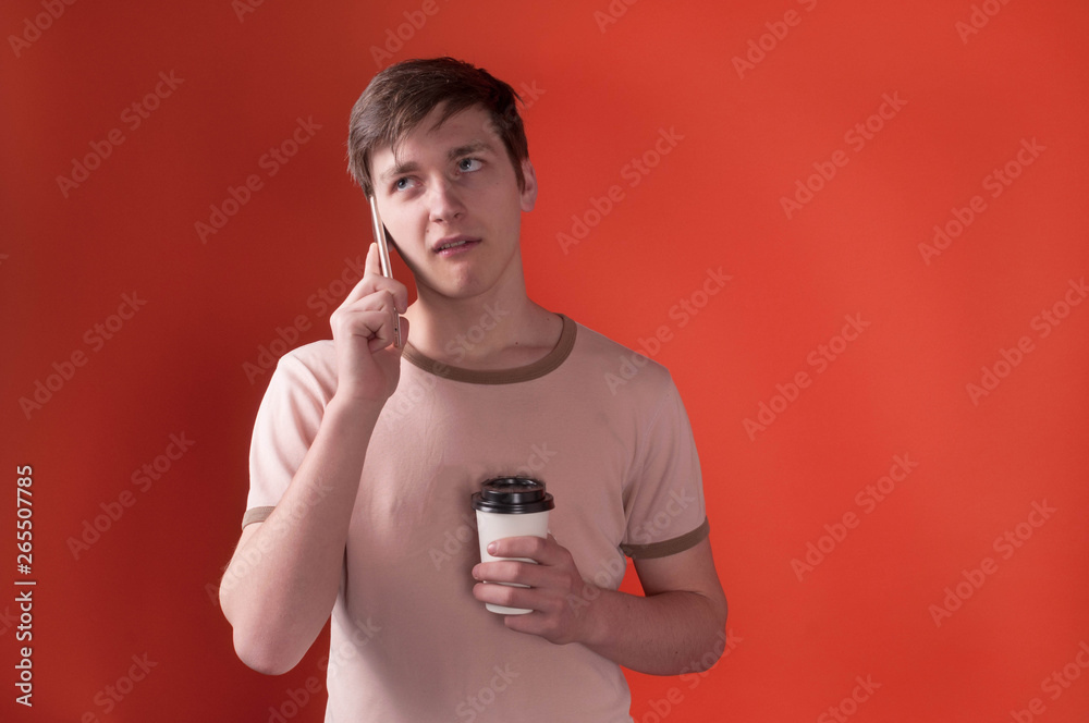 annoyed young man in beige t shirt standing and holding paper cap with coffee and talking on smartphone on orange background with copy space annoyed young man in beige t shirt standing and holding paper cap with coffee and talking on smartphone on orange background with copy space